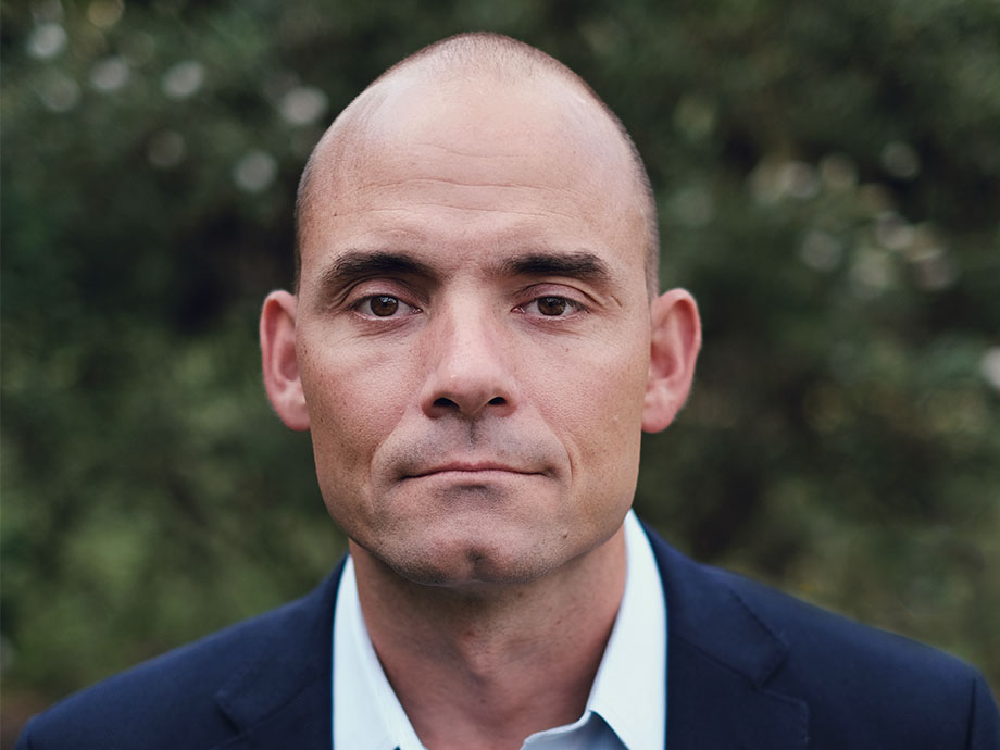Close-up outdoor headshot of a white man with a closely shaved head, wearing a dark blazer over a white collared shirt. The man is centered in the frame and facing the camera, with a softly blurred green foliage background and natural, even lighting.