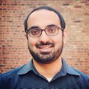 A man with dark hair and a beard wearing glasses and a blue collared shirt smiles for a portrait. A brick wall serves as the background.