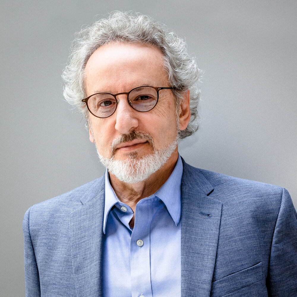 Professional portrait of a man with curly gray hair, a neatly trimmed white beard and mustache, and round eyeglasses. He is wearing a light blue dress shirt and a textured blue blazer, looking directly at the camera with a calm, confident expression. The image is framed from the chest up against a soft gray background with even studio lighting.