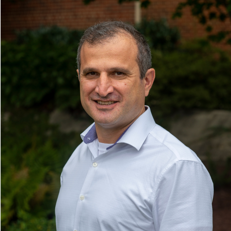 A man with dark gray hair wearing a light blue button-up shirt smiles for a portrait with foliage in the background.