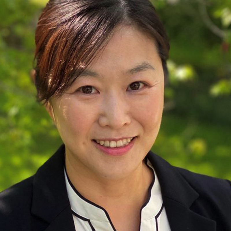 An Asian woman with dark hair smiles while wearing a black blazer over a white blouse. Blurred trees make up the background.
