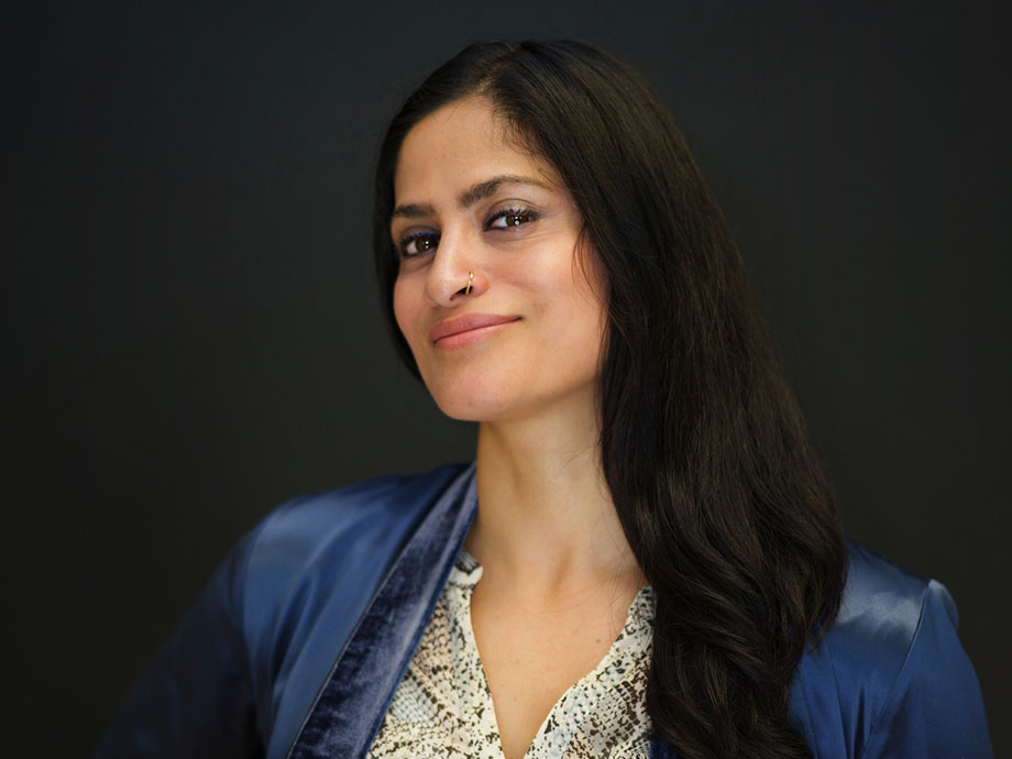 A woman with long black hair wearing a blue jacket and lacy white blouse smiles at the camera.
