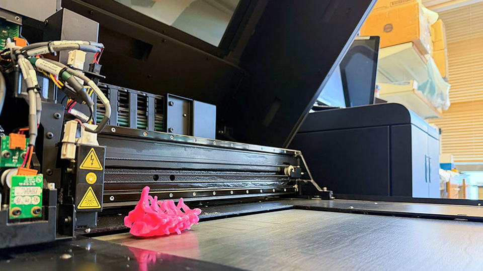 A pink 3D‑printed model of a spine resting on the build plate inside an industrial 3D printer.