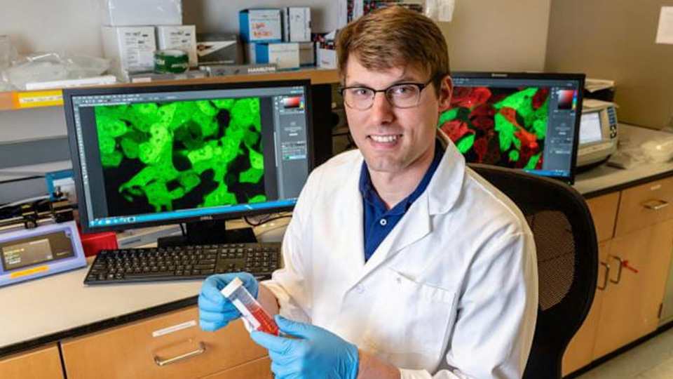 A researcher in a laboratory holds a sample vial while seated at a workstation with computer monitors displaying fluorescent microscopy images.