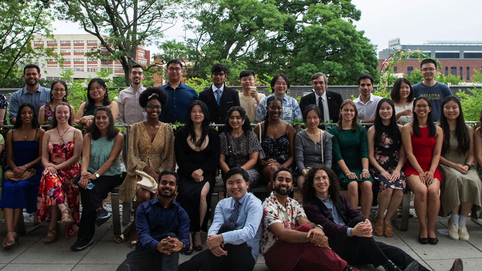 A large group of people dressed in semi-formal and casual attire posing together outdoors on a terrace with trees and campus buildings in the background.