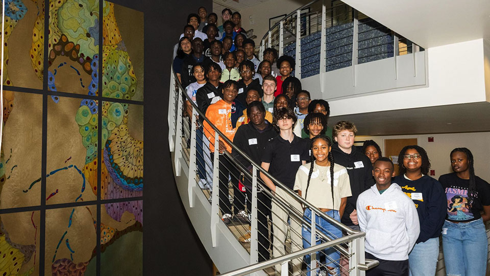 A large group of students standing together on a staircase in a building, posing for a group photo next to a colorful wall mural.