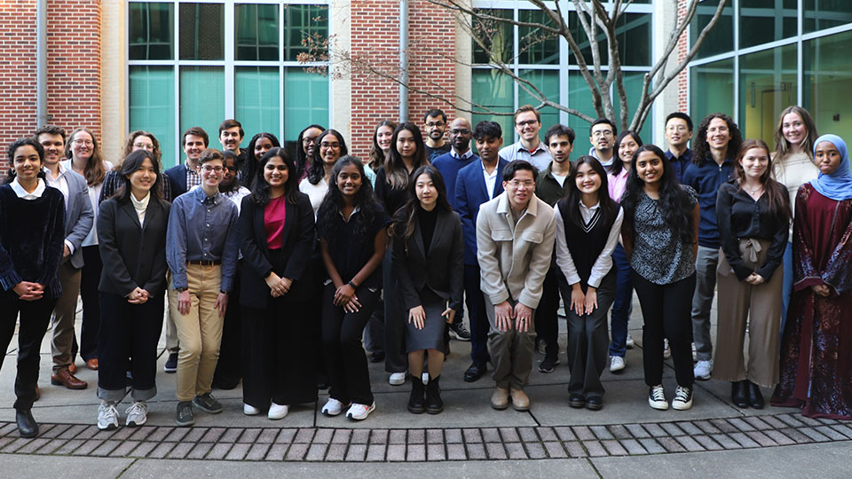 A large group of students standing together outdoors in a courtyard, posing for a group photo in front of a building with large windows and brick columns.