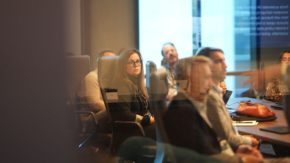 A group of professionals seated around a conference table during a meeting, listening and taking notes in a modern office setting.