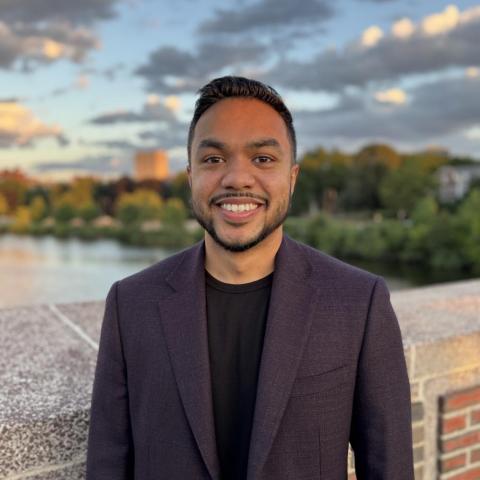 A man with dark hair and a short beard smiles for a portrait. He wears a dark purple blazer over a black shirt and stands on a bridge with water and a sunset in the background.