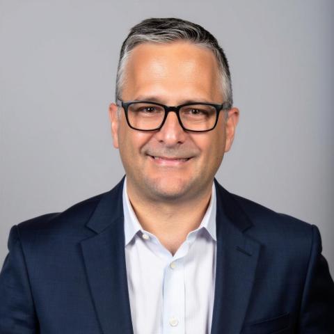 A man with gray hair and black glasses wearing a button-up shirt under a navy blazer