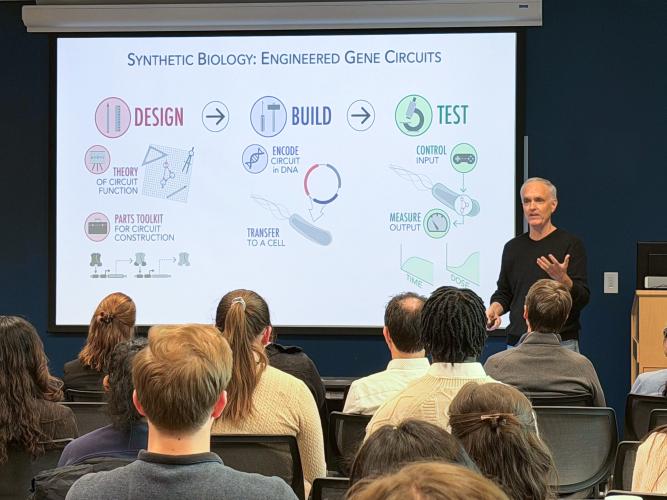 A presenter stands at the front of a lecture room speaking to a seated audience while a projected slide titled “Synthetic Biology: Engineered Gene Circuits” illustrates the design–build–test cycle with diagrams and icons explaining gene circuit construction and testing.