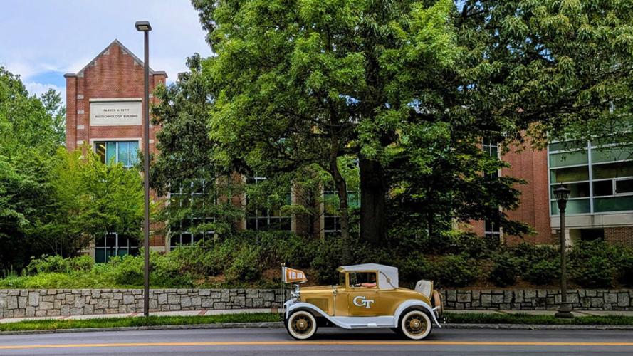 The Ramblin Wreck drives past the Petit Biotechnology Building, a red brick building with large trees in the foreground.