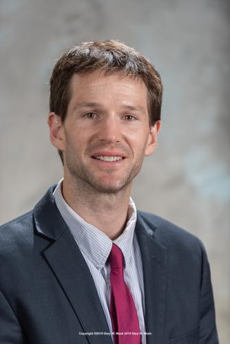 A man with brown hair and a short beard smiles for a portrait while wearing a dark blue suit and red tie.