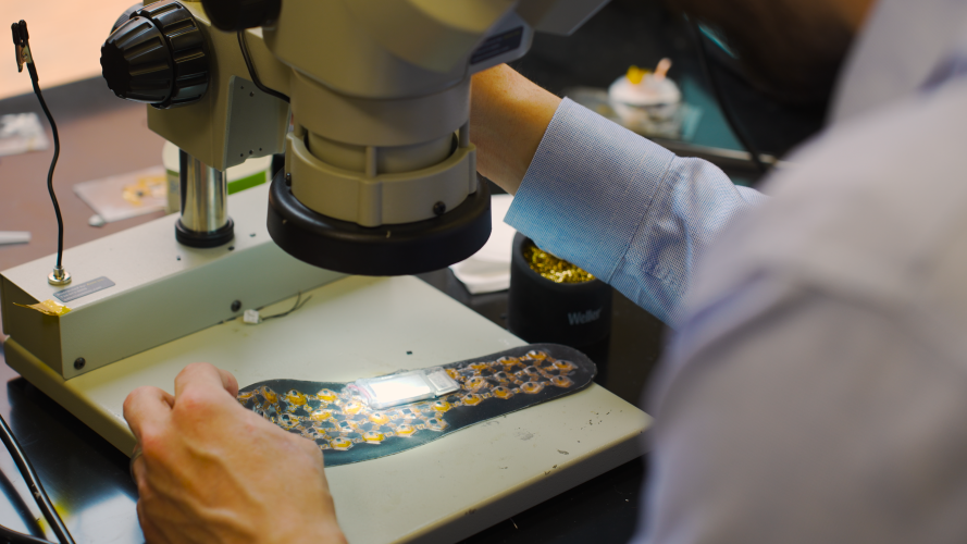 Close-up of hands positioning a flexible haptic device with embedded electronics under a microscope, highlighting the small components and patterned array used to deliver sensory feedback.