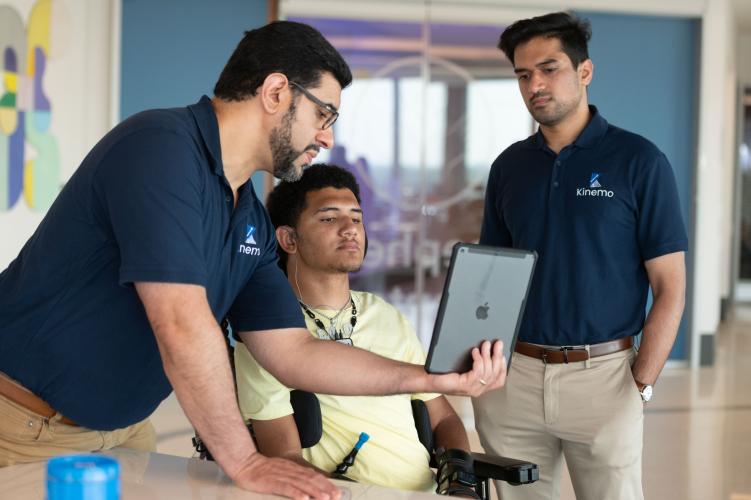 Three people review a tablet together in a lab setting, with one seated individual using a powered wheelchair and assistive device.