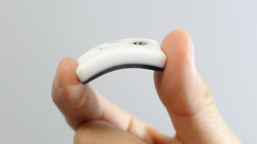 Close-up of a flexible white wearable sensor device held between two fingers, showing its curved shape against a plain light-gray background.
