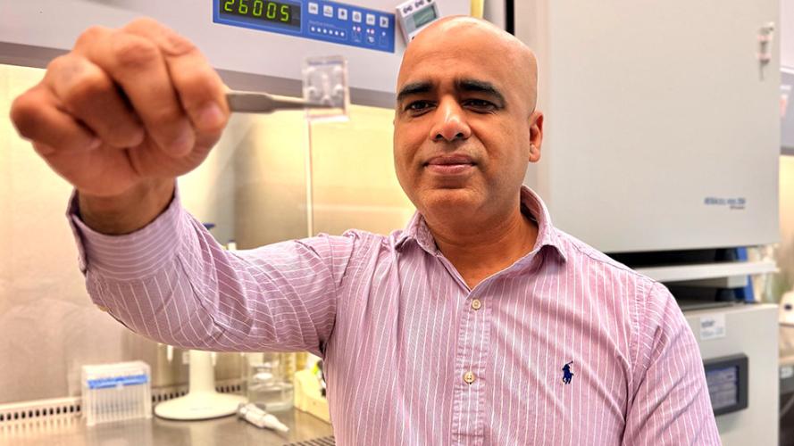A man sits in a lab in front of a fume hood and uses tweezers to hold a plastic chip out toward the camera.