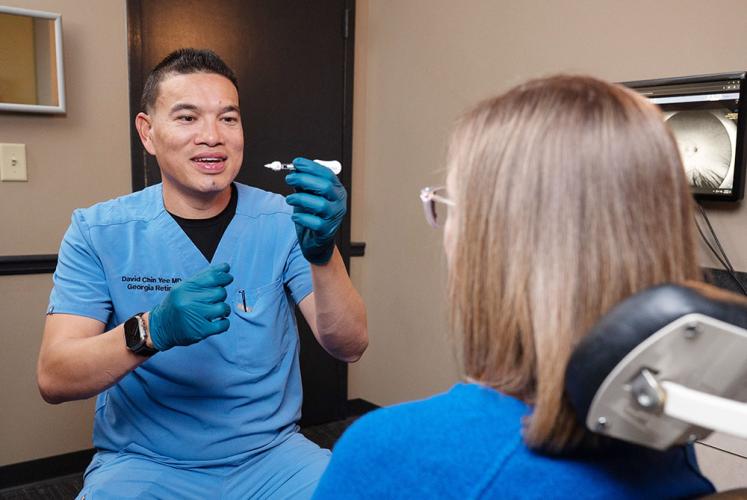 Person in blue medical scrubs demonstrates a small device to a seated patient in a clinical exam room with medical equipment visible in the background.